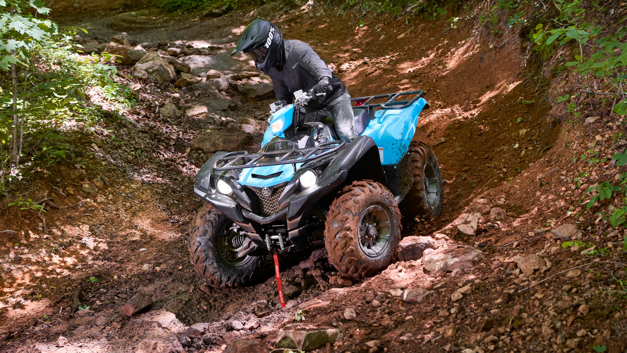 Helmeted rider on a blue quad crossing a rocky forest trail